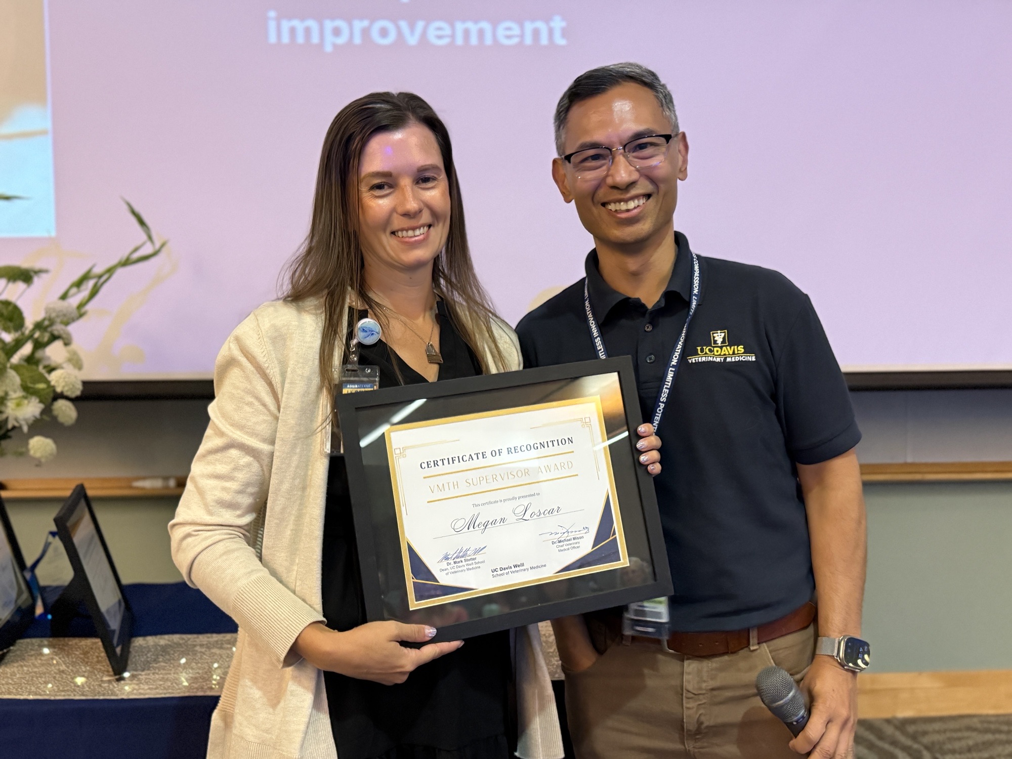 Two smiling colleagues holding a framed award in a conference room, candid photo