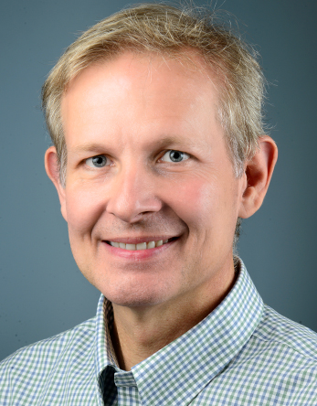 Smiling headshot photo of a middle-aged man with light hair, checkered shirt, gray background