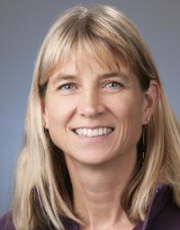 Smiling headshot of a middle-aged blonde woman wearing a purple blazer, gray background.